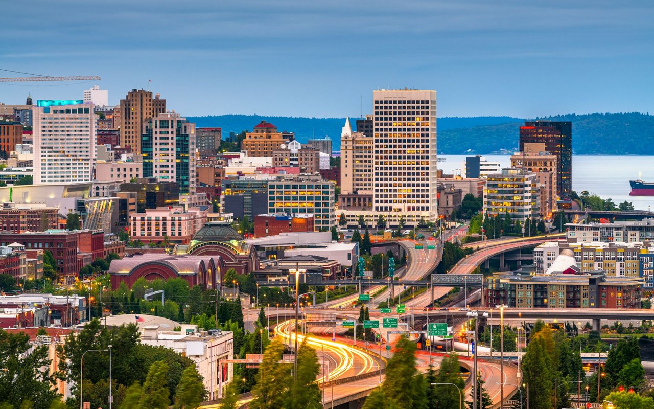 Tacoma skyline above freeway light trails, with waterfront and wooded hills beyond.