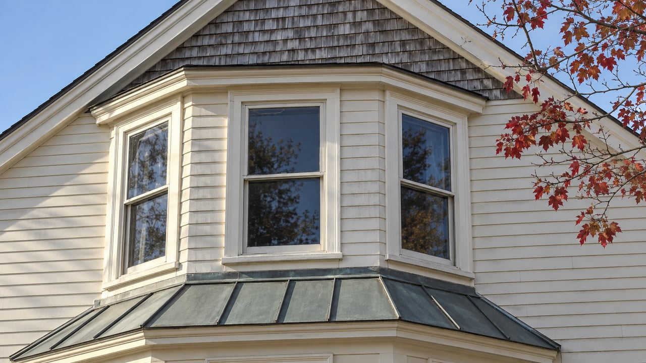 Late-fall sunlight on an ivory clapboard house's bay windows, weathered cedar gable, and gray-green metal roof beside a red maple branch.