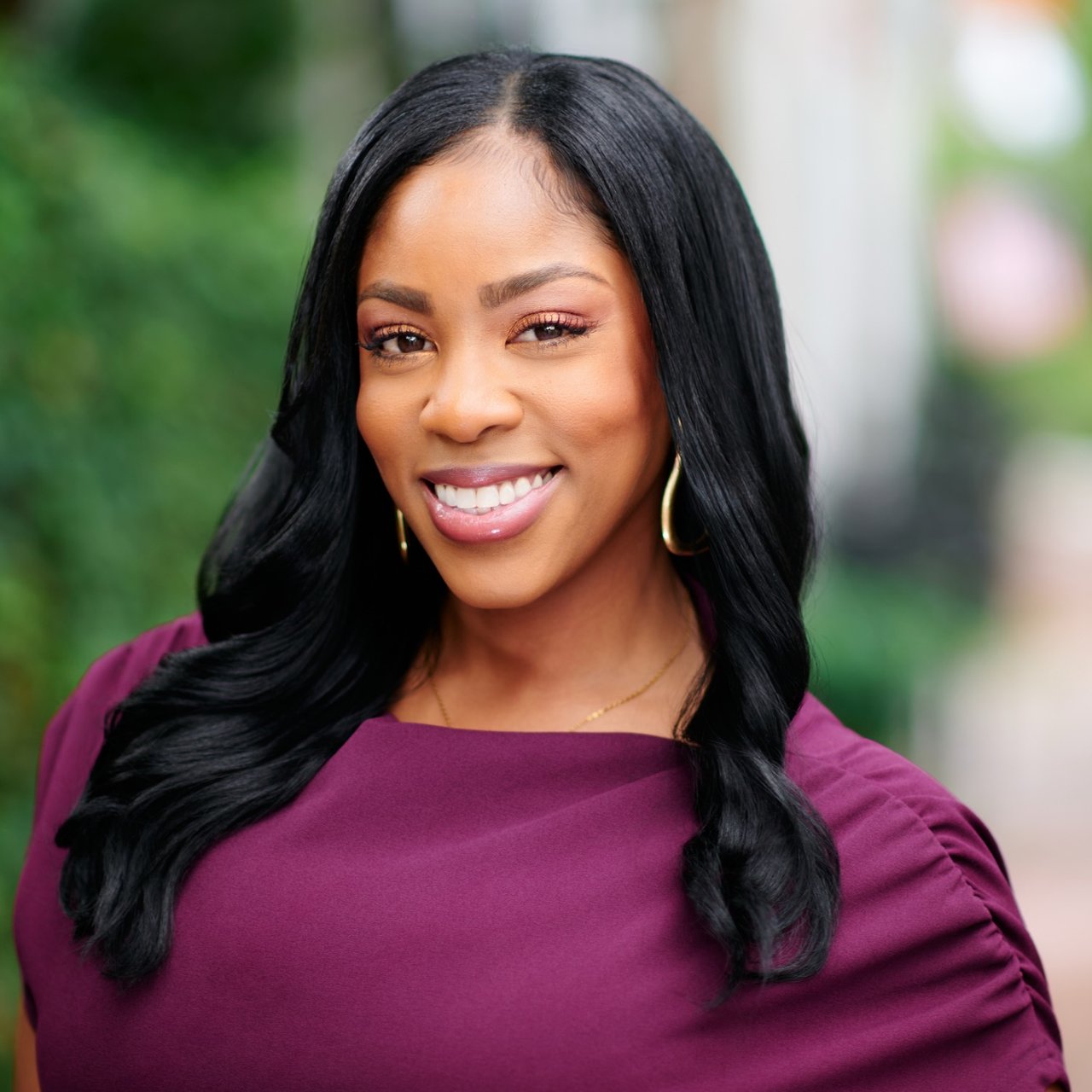Tiffanie Cross smiling in a plum blouse, with long black hair and gold hoop earrings.