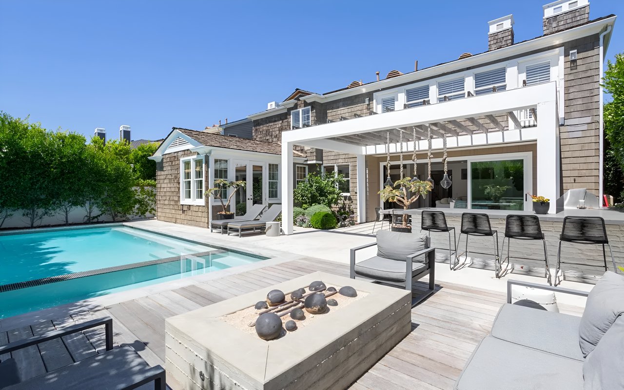 A sunny backyard with a large swimming pool, a modern fire pit, an outdoor kitchen, and a patio with seating areas.