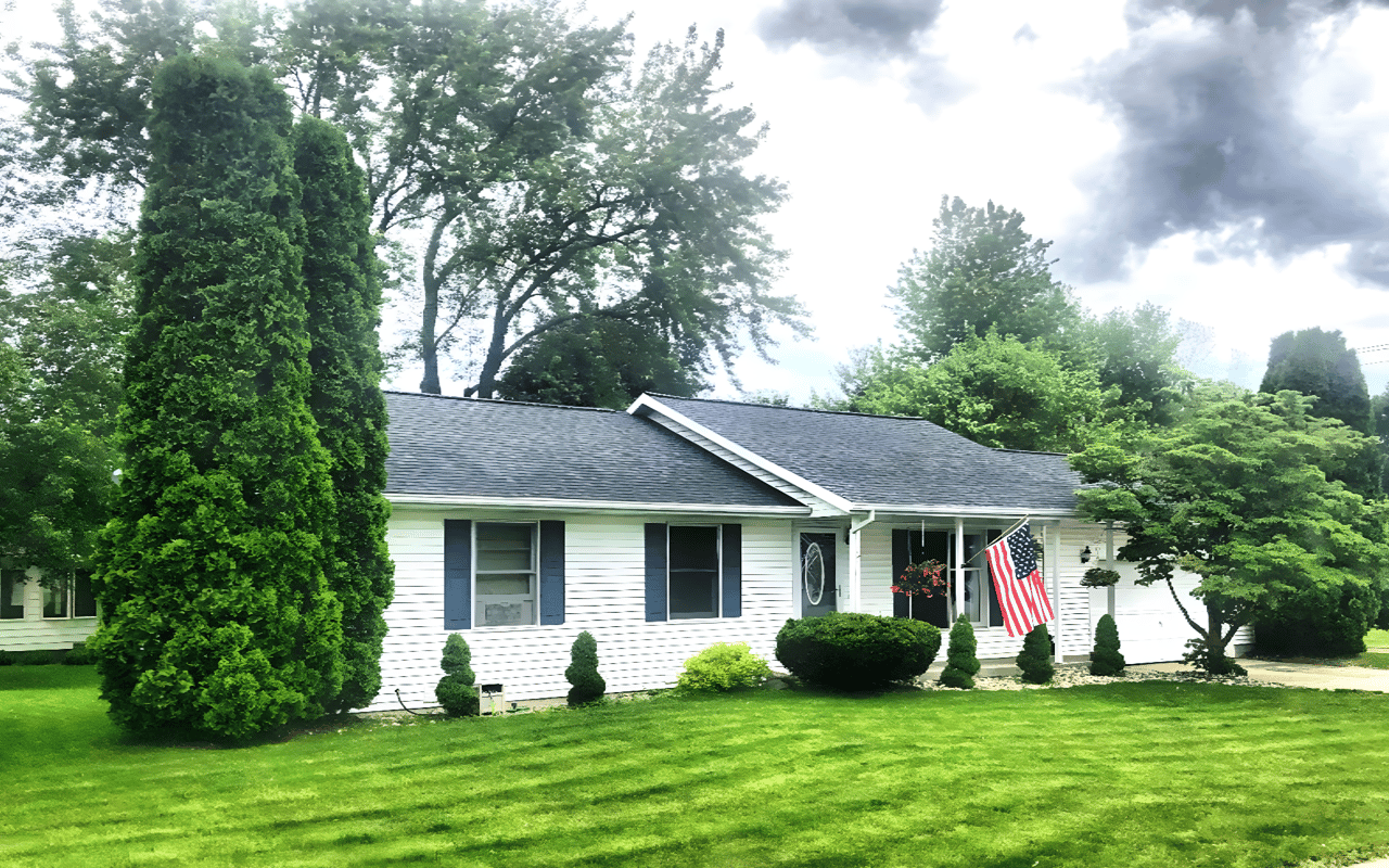White single-story house with blue shutters, an American flag, and a landscaped lawn.
