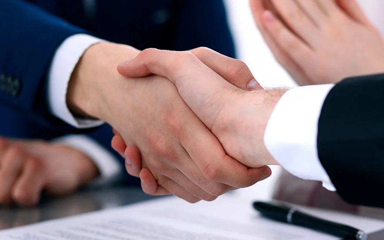 Two people shake hands over paperwork, with a pen on the table.
