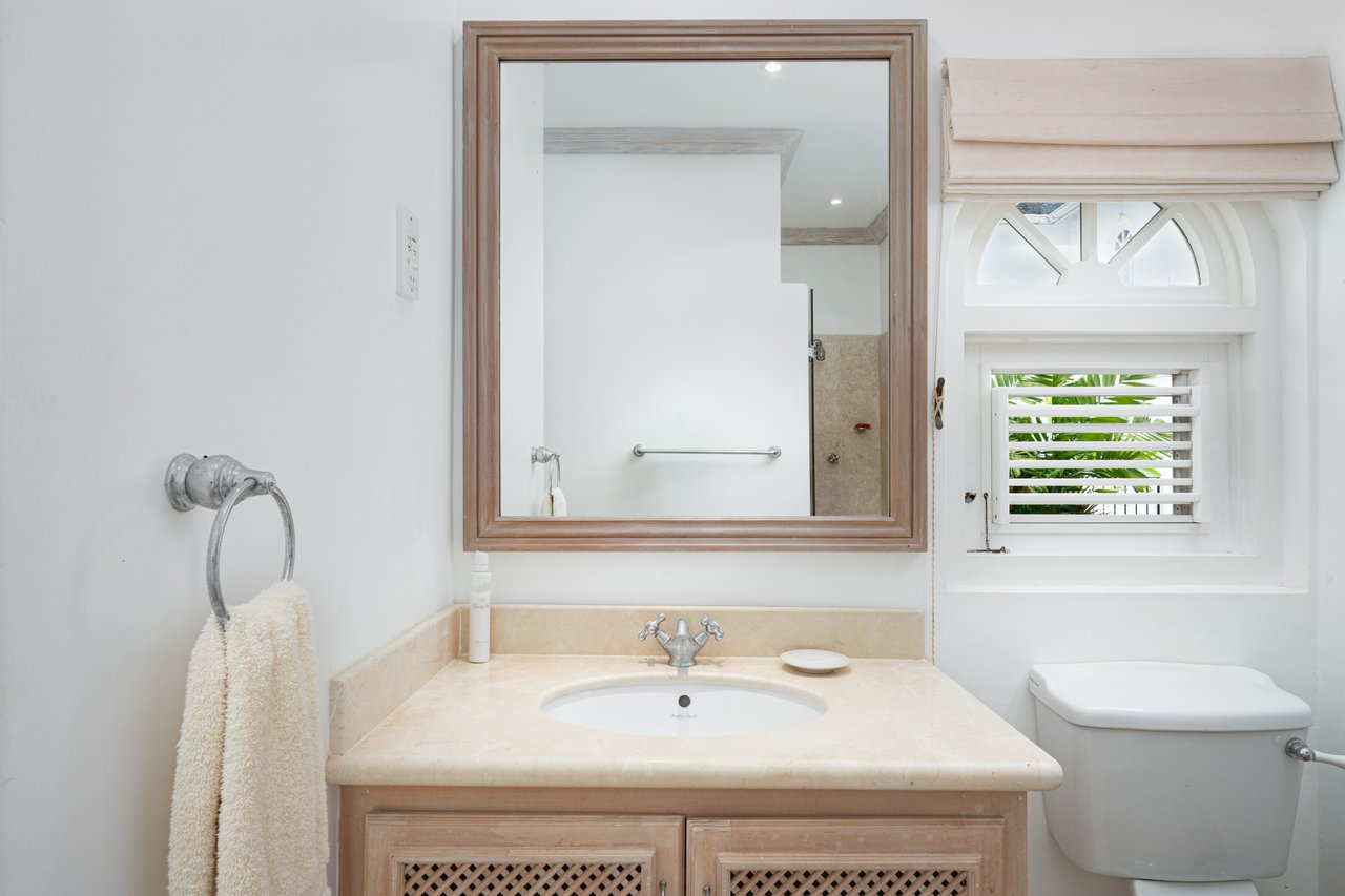 Bathroom with a pale stone vanity, wood-framed mirror, and shuttered window overlooking greenery.