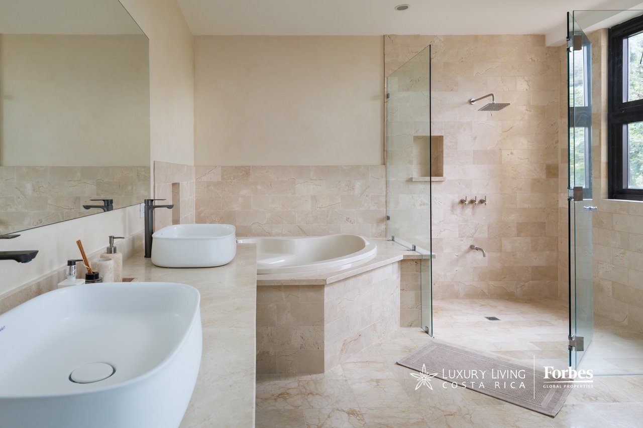 Penthouse bathroom with beige stone tile, two vessel sinks, soaking tub, and glass-enclosed shower.