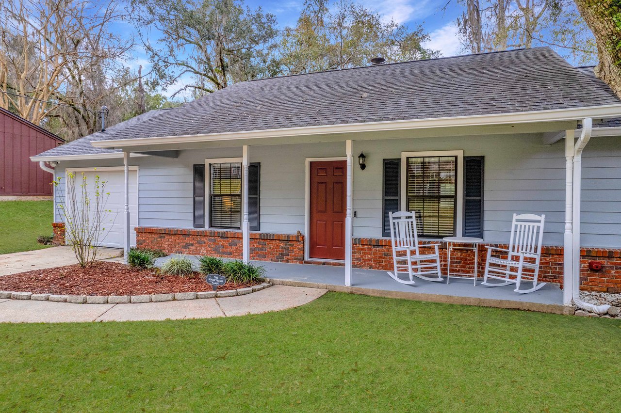 Single-story house with light blue siding and a brick foundation. Features a porch with two white rocking chairs, red door, and lush green lawn. Cozy and inviting atmosphere.