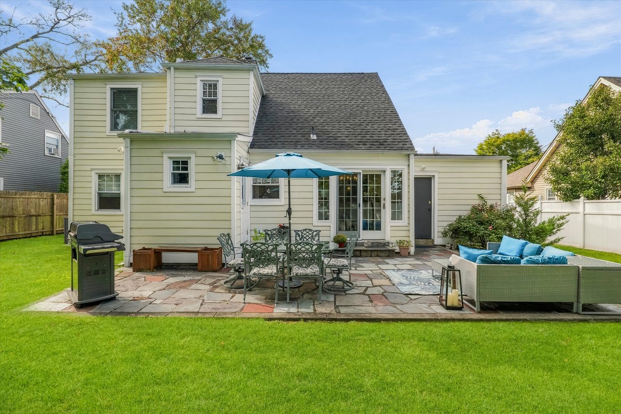 Home’s rear patio with outdoor dining under a blue umbrella and lounge seating beside the lawn.