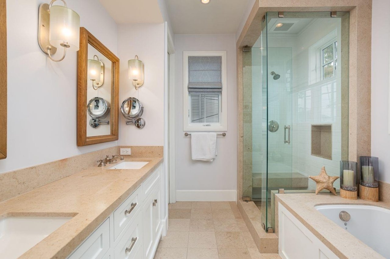 A large, bright bathroom with a double vanity, glass-enclosed shower, bathtub, and light-colored tiled floors and countertops.