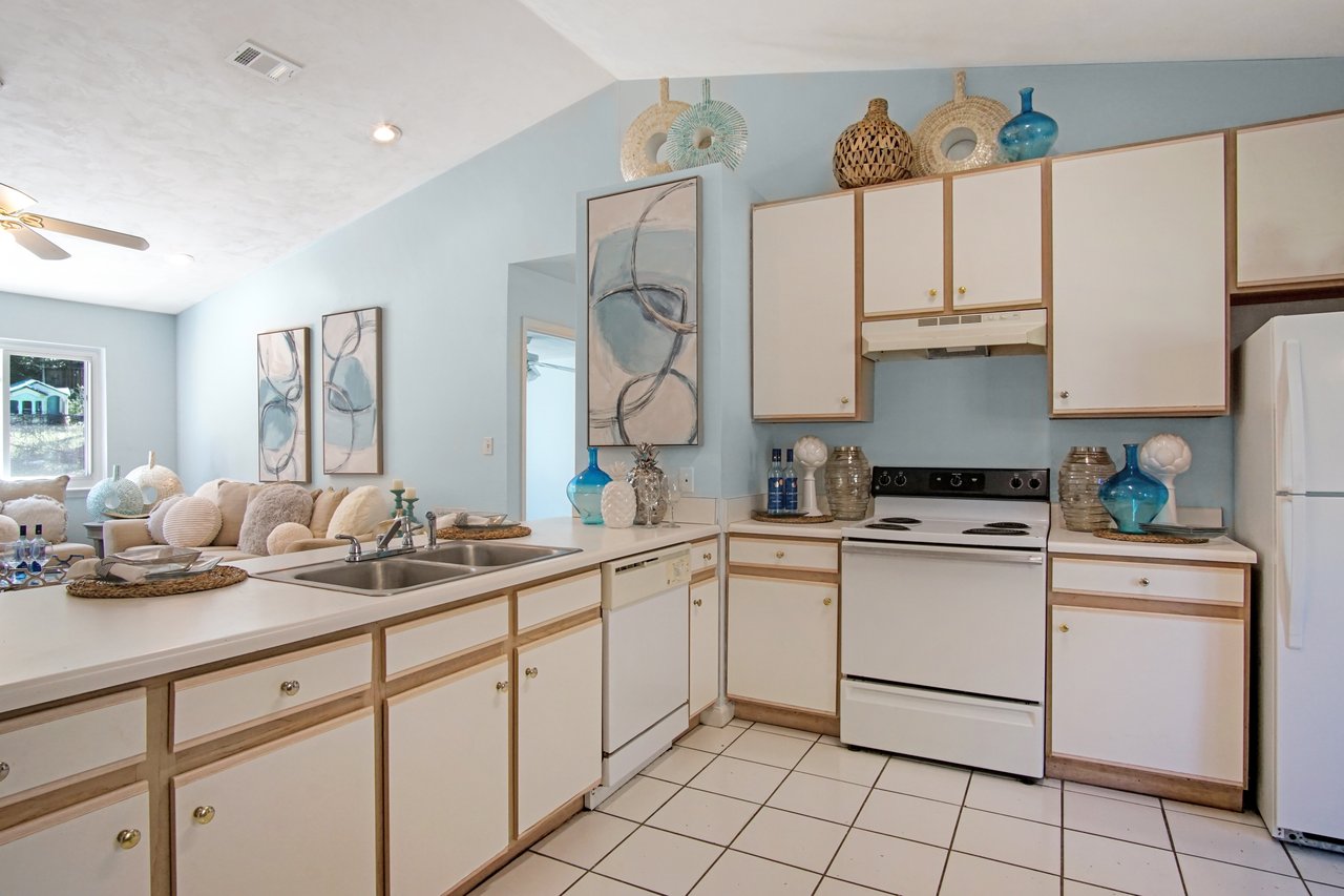 Bright kitchen with blue walls, white cabinets, and appliances. Decor includes abstract artwork, wicker baskets, and blue vases, conveying a coastal vibe.