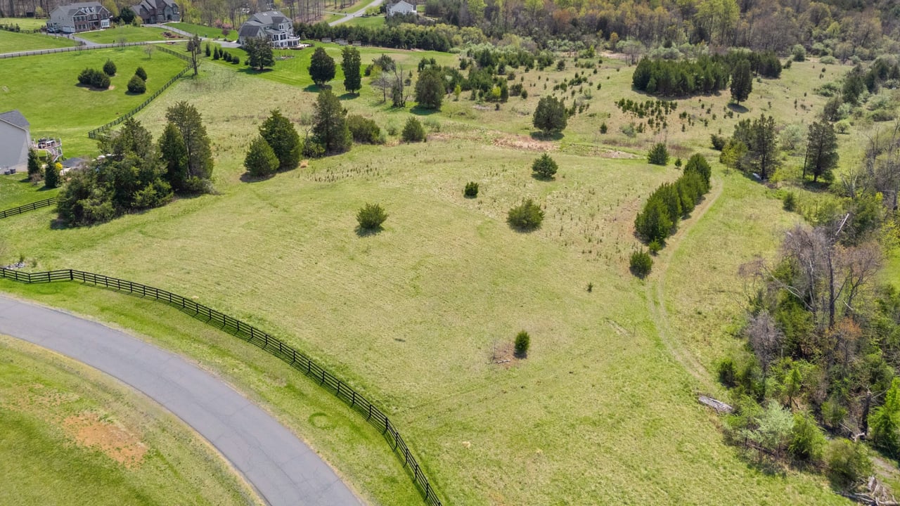 Aerial pasture with black fencing beside a curving road and neighboring homes in the distance.
