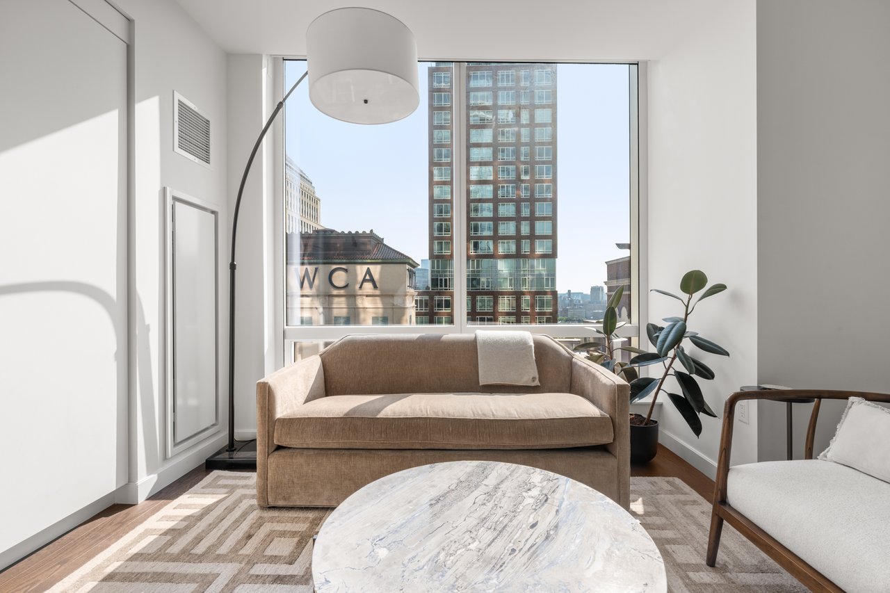 Furnished living room with a tan sofa and large windows overlooking city buildings.