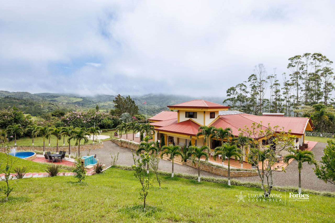 Mountain house with red roofs, a small pool, palm trees, and cloud-covered green hills.