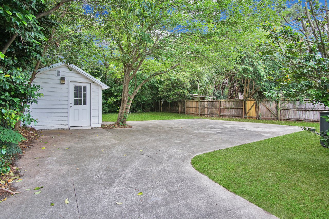 A cozy white shed with a glass-paneled door sits beside a concrete driveway, bordered by lush trees and a wooden fence, evoking a serene, inviting atmosphere.