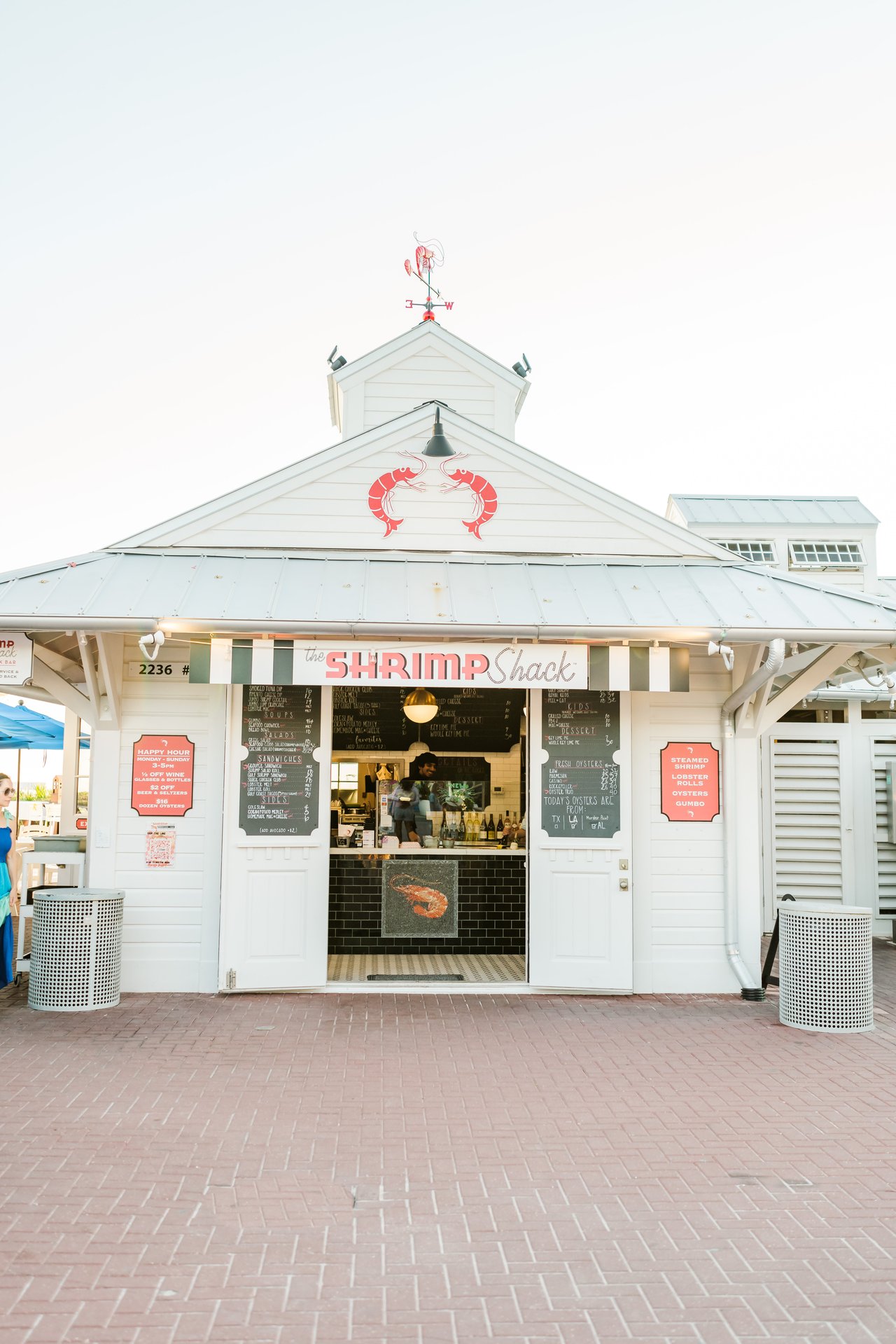 The white open-air 'Shrimp Shack' kiosk with a shrimp weathervane and chalkboard menus on a herringbone brick plaza in Seaside on 30A, Florida