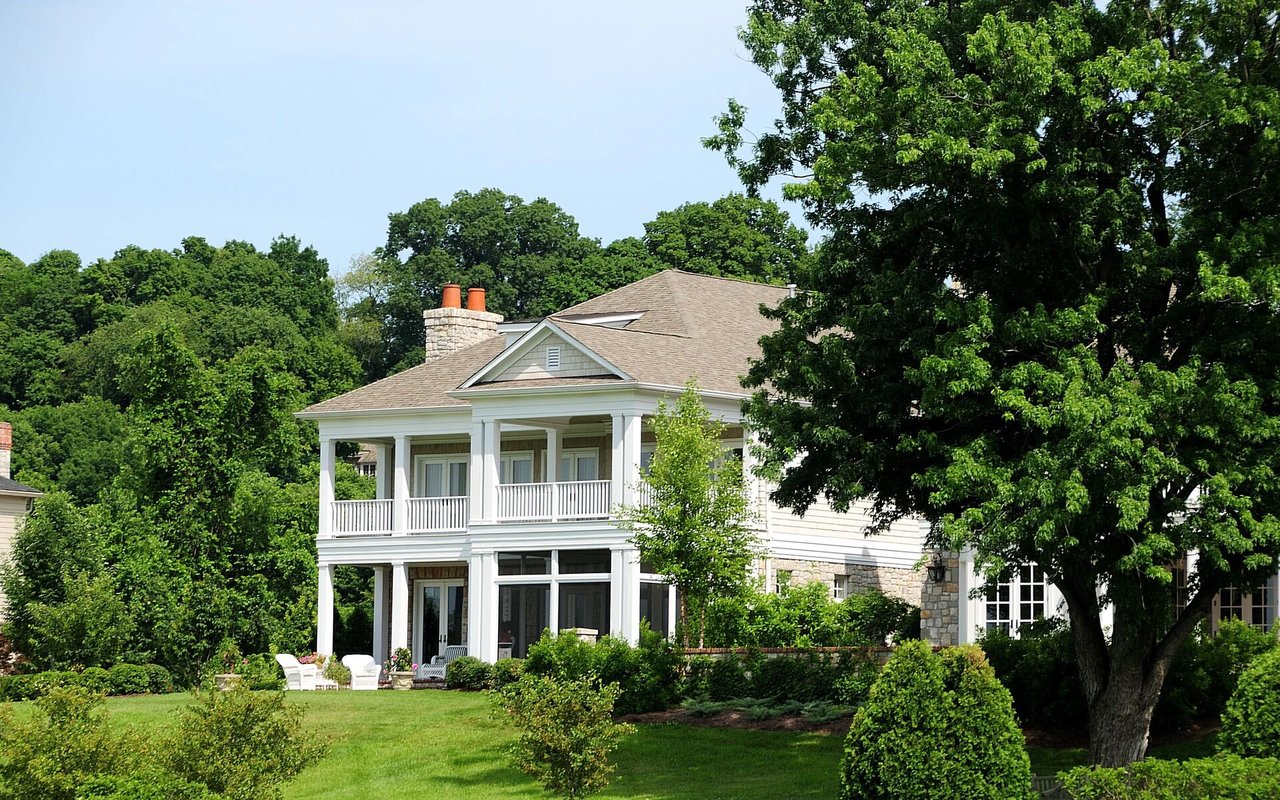White two-story house with covered porches and stone accents, surrounded by mature trees and lawn.
