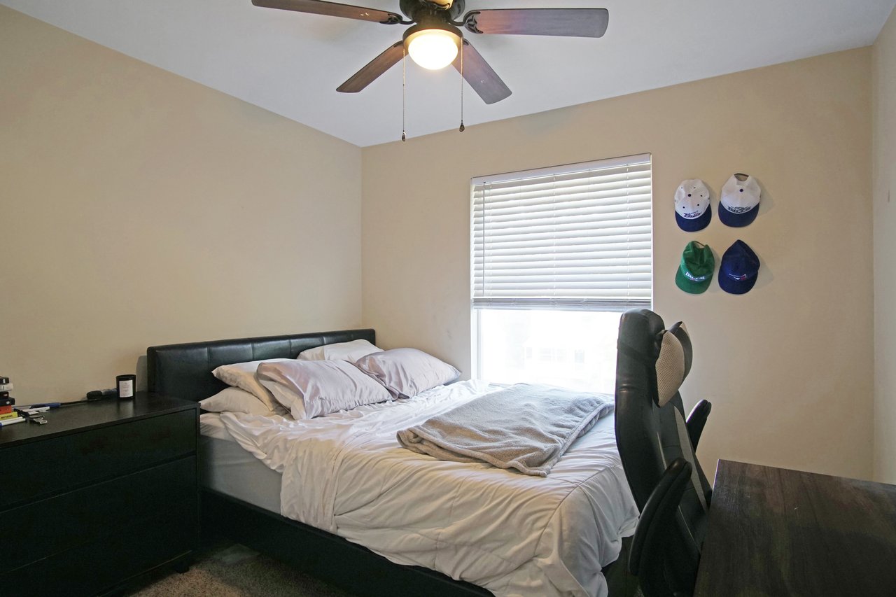 Bedroom at 529 Collinsford Road, Tallahassee, Florida, featuring a bed with white bedding and gray pillows, a ceiling fan with light fixture, and a window with blinds letting in natural light. The room includes a dark dresser, desk with chair, and wall-mounted baseball caps for decoration.