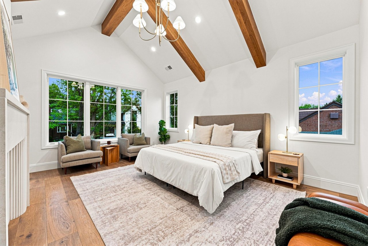 Staged bedroom with a vaulted ceiling, exposed wood beams, and a seating area beside large windows.