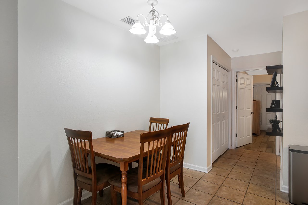 Cozy dining area with a wooden table and four chairs under a bright chandelier. The room has white walls, tile flooring, and a hallway with shelves.