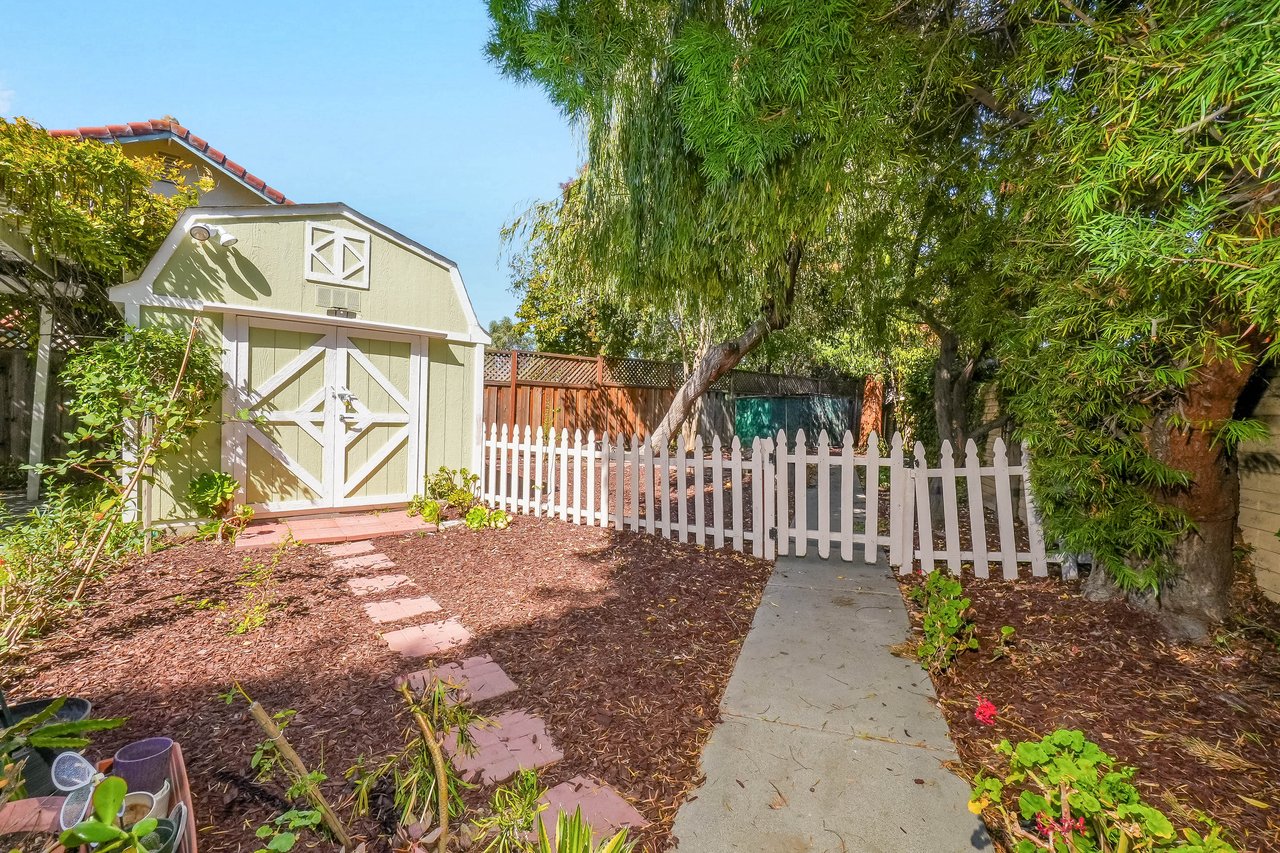Backyard with a pale green shed, white picket fence, and walkway beneath mature trees.