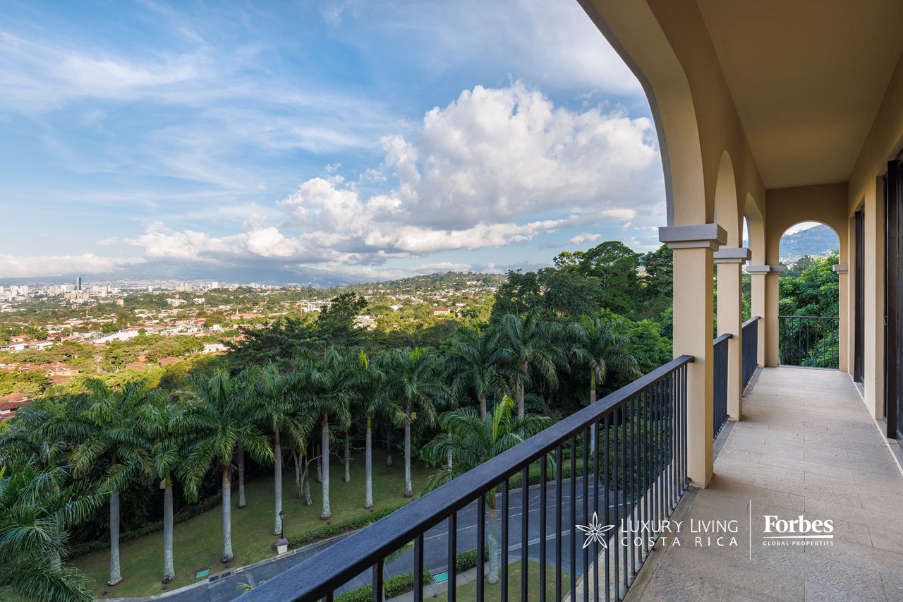 Covered balcony overlooking a green canopy, city, and distant mountains.