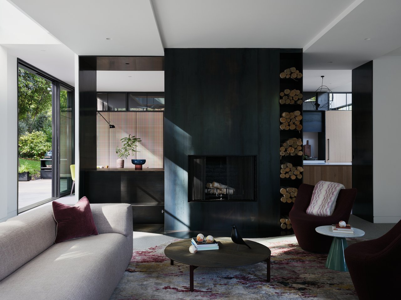 Modern living room with a dark fireplace wall, built-in firewood niche, and garden-facing windows.