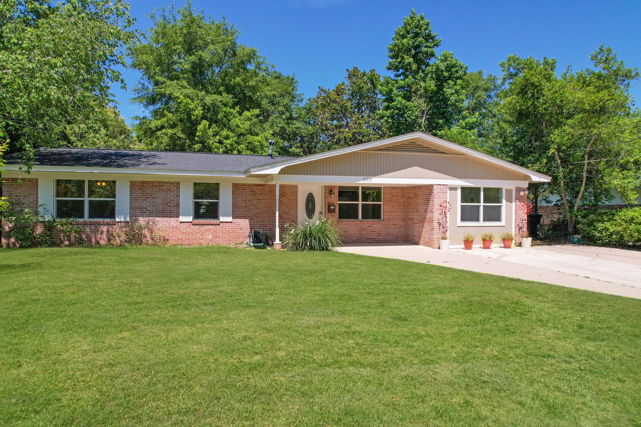 Single-story brick house with a dark roof surrounded by lush green trees. The front yard has well-kept grass, and there are potted plants by the entrance.