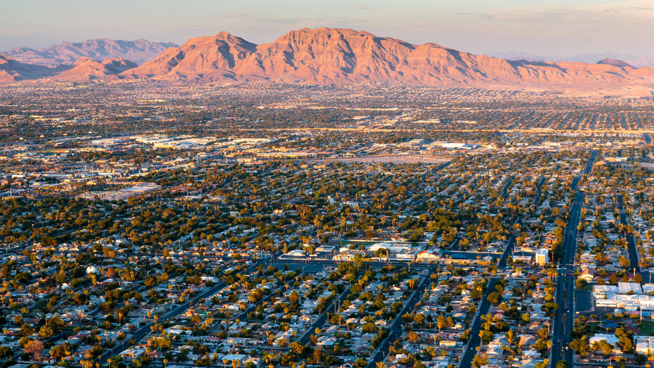 Aerial view of Las Vegas Valley neighborhoods and streets with desert mountains illuminated by warm sunlight in the background.