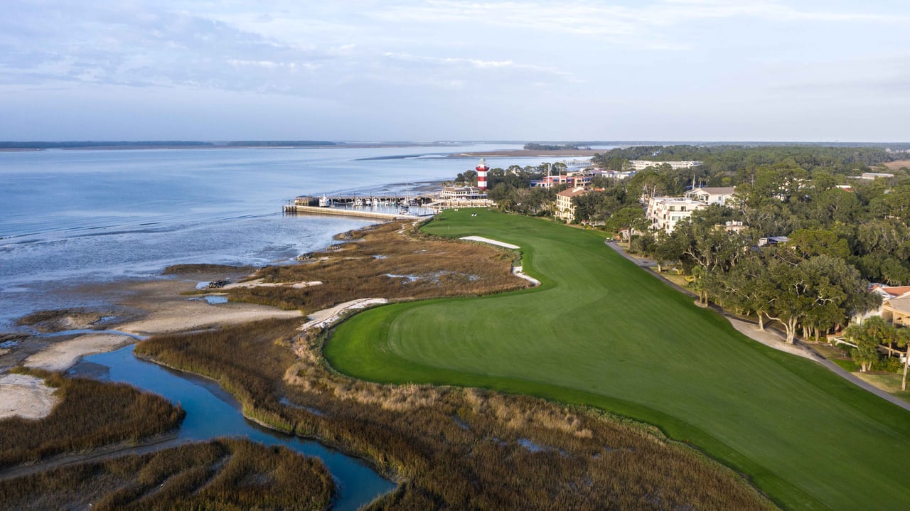 Coastal golf course beside marshland and water, with a red-and-white lighthouse in the distance.