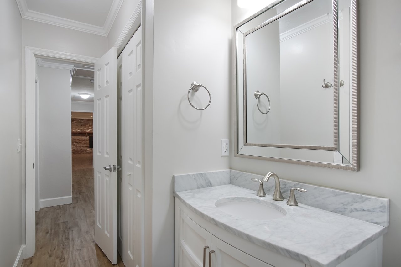 A small bathroom with a light-colored marble countertop, sleek silver faucet, and large rectangular mirror. Neutral walls and wood floor evoke a clean, modern feel.
