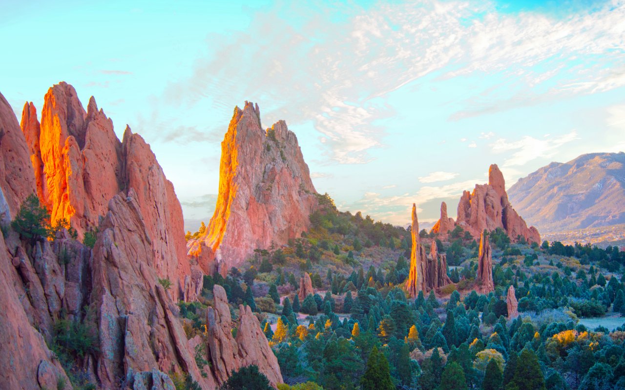 Garden of the Gods’ red sandstone formations rise above forested foothills in warm evening light.