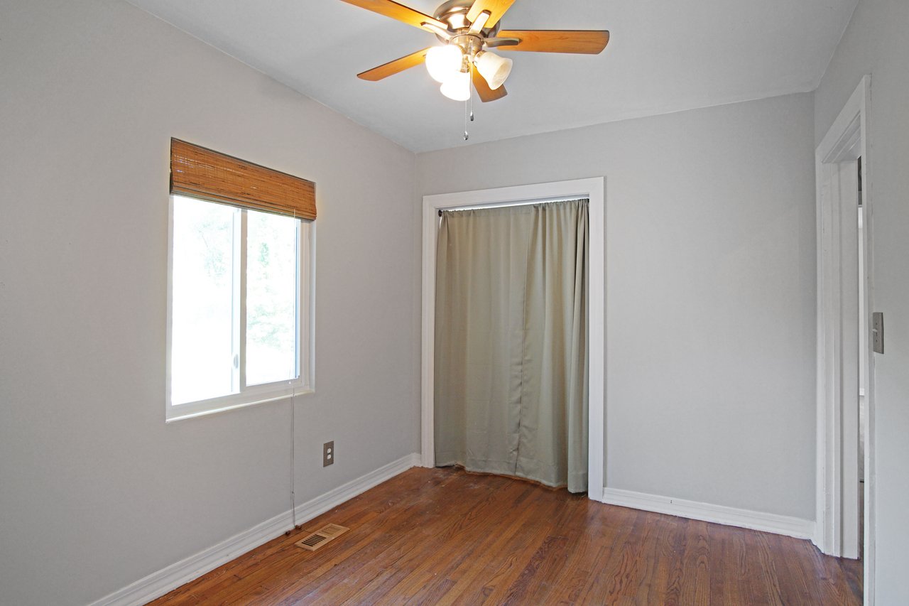 Minimalist room with wooden flooring and a ceiling fan, featuring a bright window with bamboo blinds and a curtained closet, exuding a simple, airy vibe.