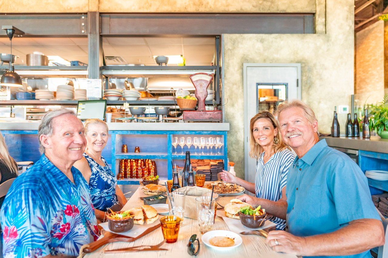 Four adults sharing a meal at a restaurant table beside an open kitchen.
