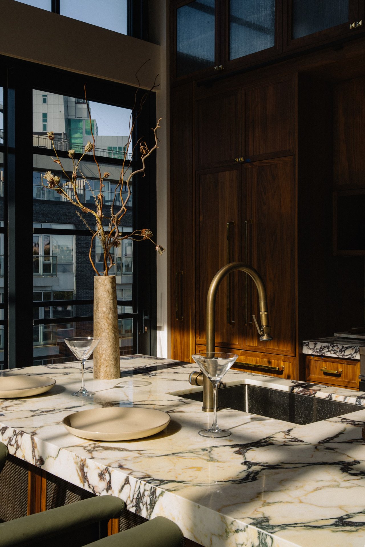 Kitchen with a veined marble island, walnut cabinetry, brass faucet, and large windows overlooking neighboring buildings.
