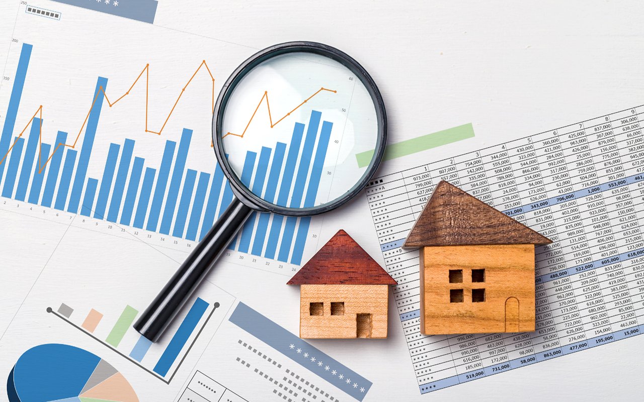 Wooden house models on housing market charts, viewed through a magnifying glass.