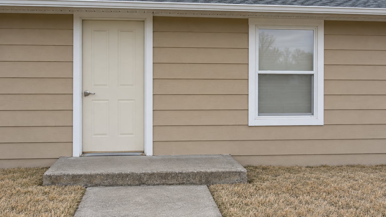 A modest tan-sided home's closed entry door and white-trimmed window above a weathered concrete stoop and dormant lawn.