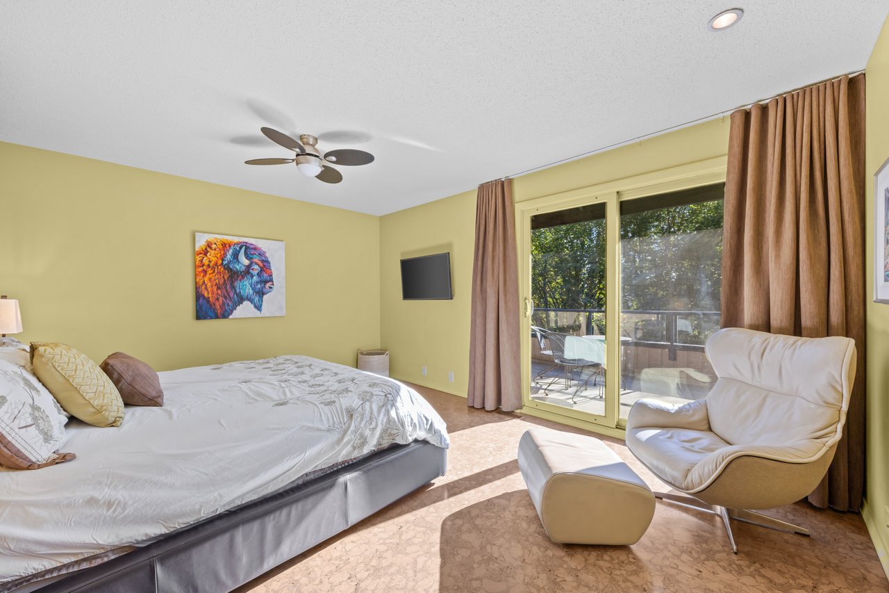 Bedroom with a large bed, white lounge chair, bison artwork, and sliding doors to a tree-lined deck.