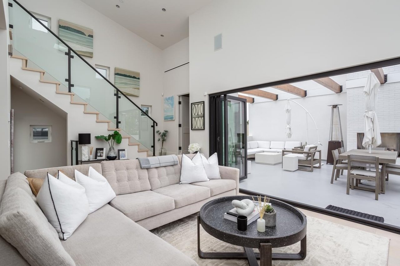 A bright, modern living room with a large sectional sofa, a staircase with a glass railing, and bi-fold doors leading to a patio.