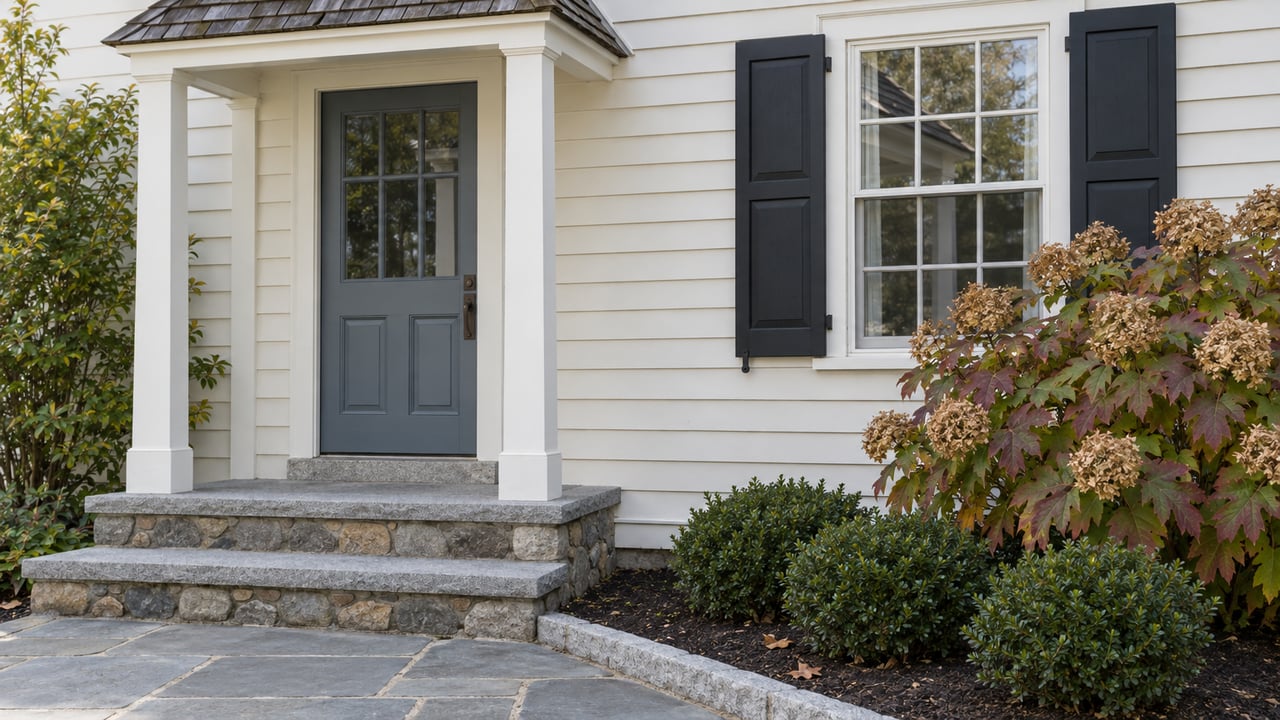 Ivory clapboard entry with a blue-gray door, paired black window shutters, granite steps and autumn oakleaf hydrangea.
