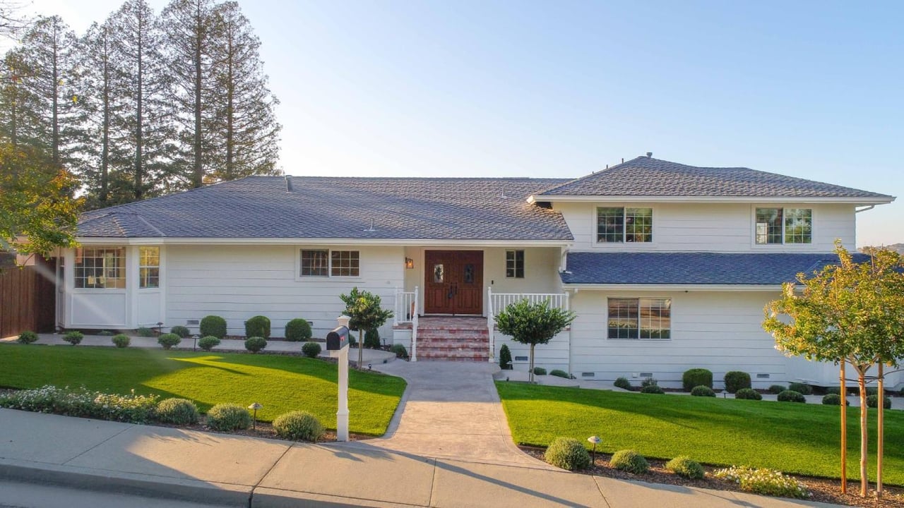 White two-story house with wooden double doors, landscaped lawn, and tall trees behind it.