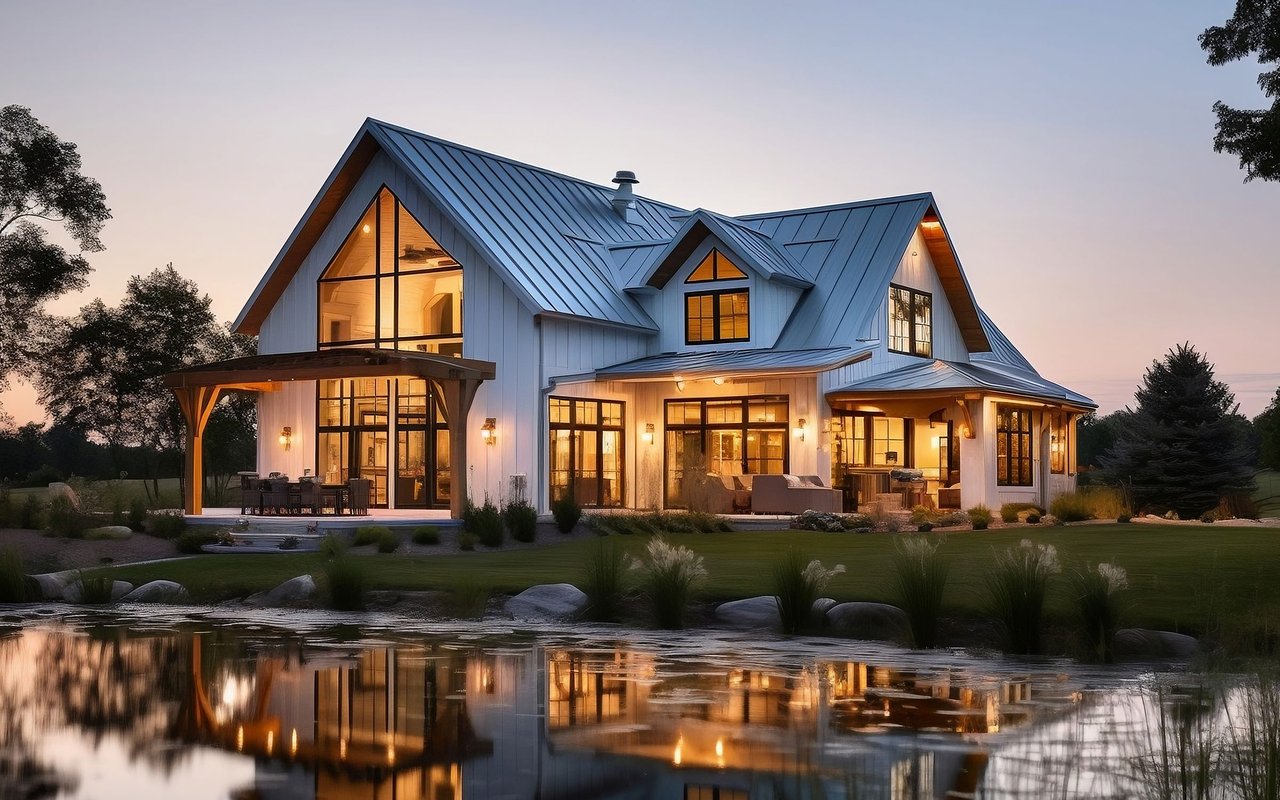 Modern farmhouse with a blue metal roof and warmly lit windows reflected in a pond at dusk.