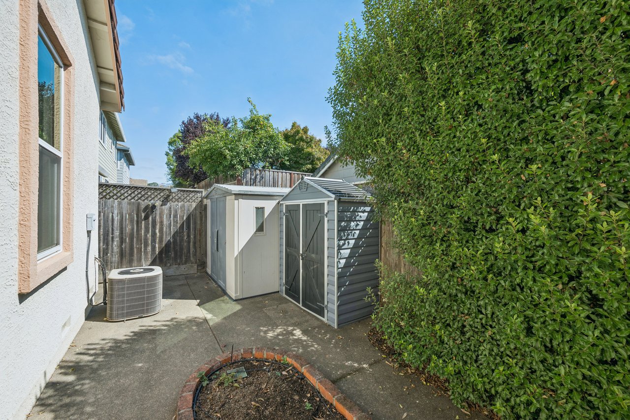 Backyard patio with a gray storage shed, air-conditioning unit, and tall hedge.