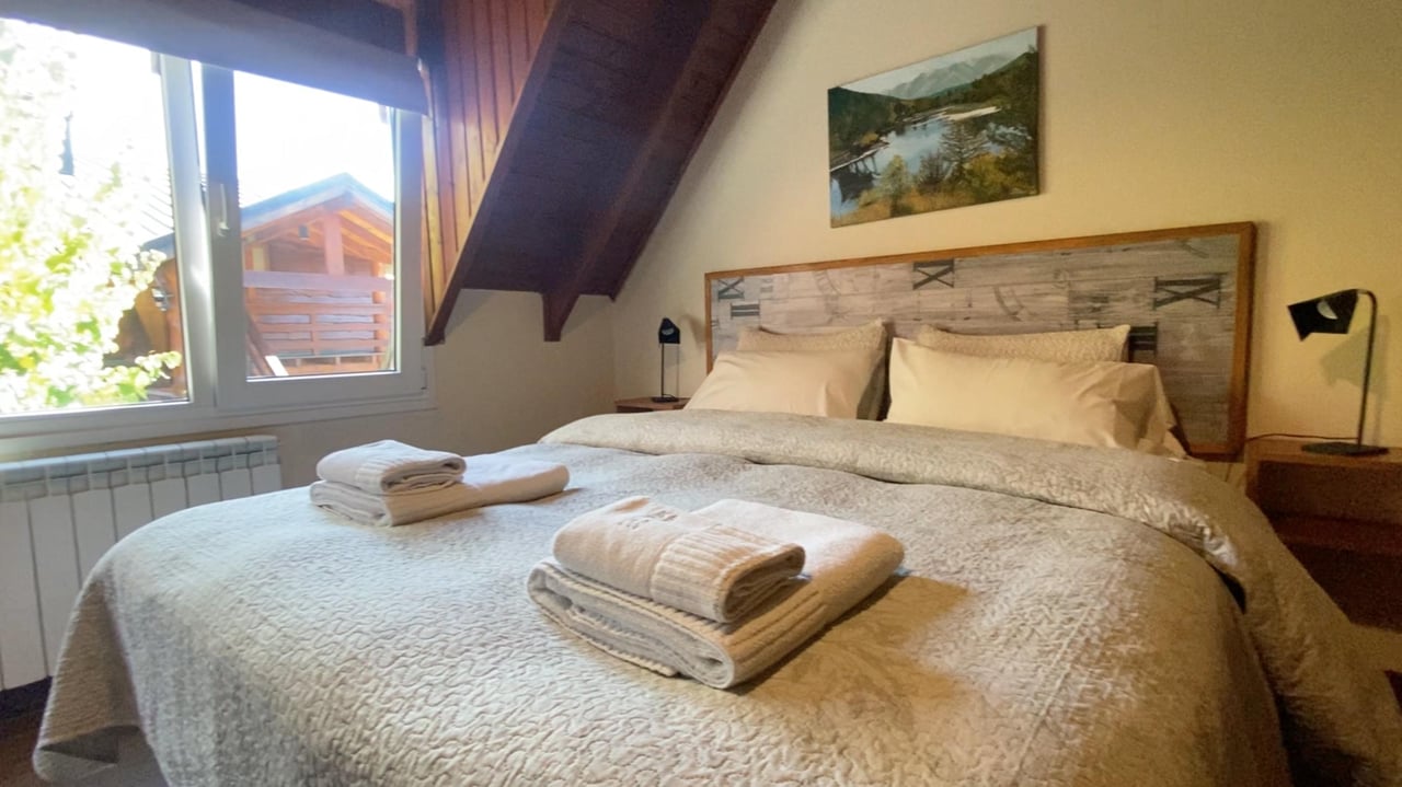 Attic bedroom with a large bed, exposed timber beams, and a window overlooking a wooden chalet.