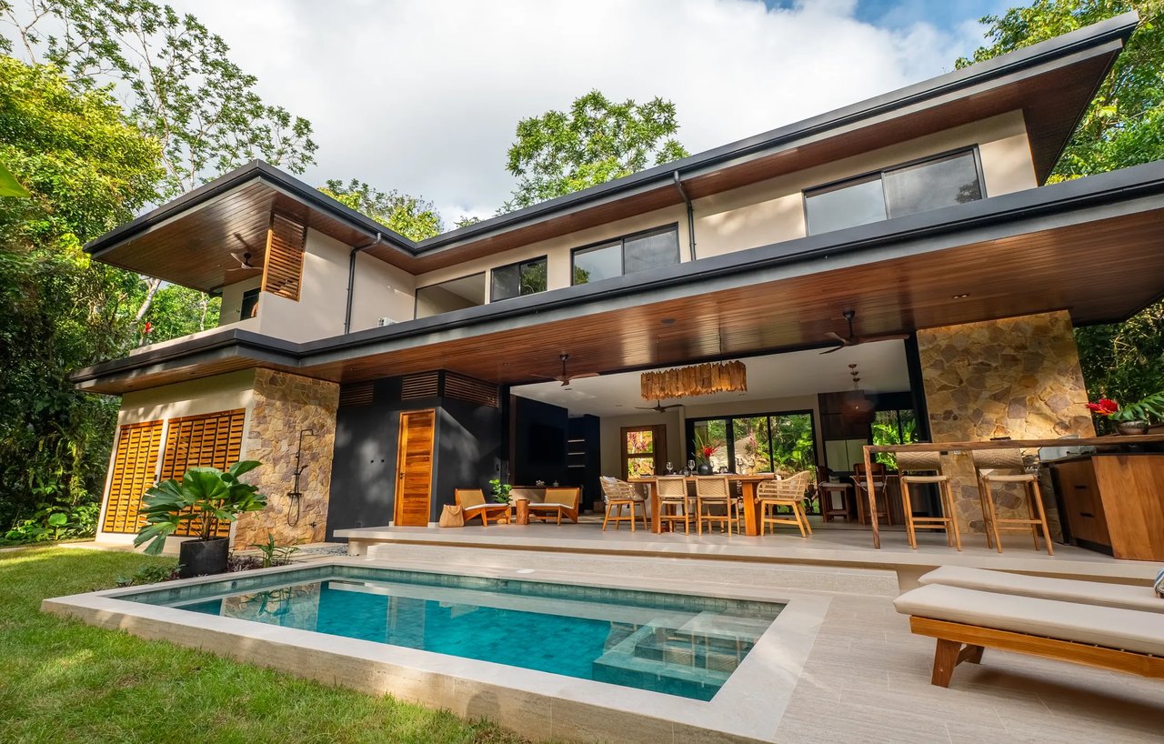 Modern two-story home with a swimming pool and covered outdoor dining area amid tropical greenery.