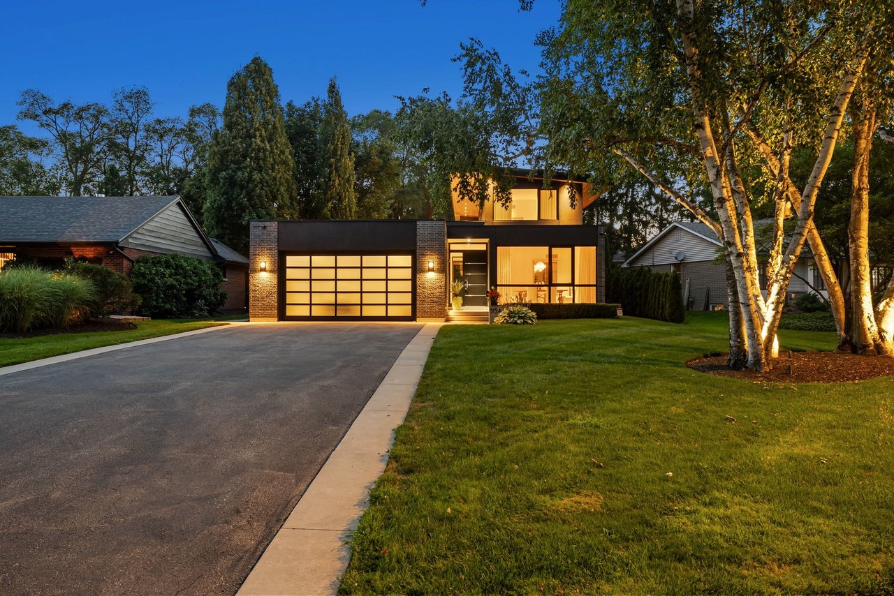 Modern glass-fronted house with illuminated windows, double garage, and broad driveway at dusk.