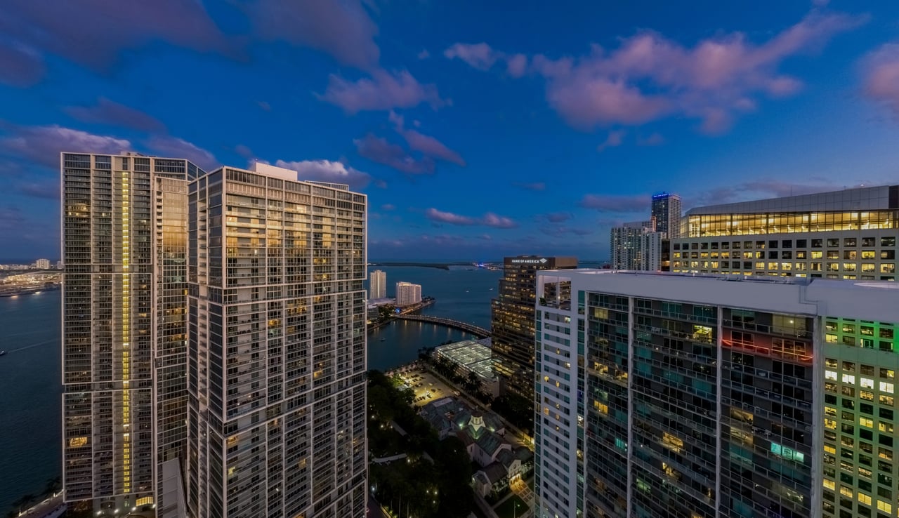 Brickell high-rises beside the bay at dusk, with illuminated windows and water between the buildings.