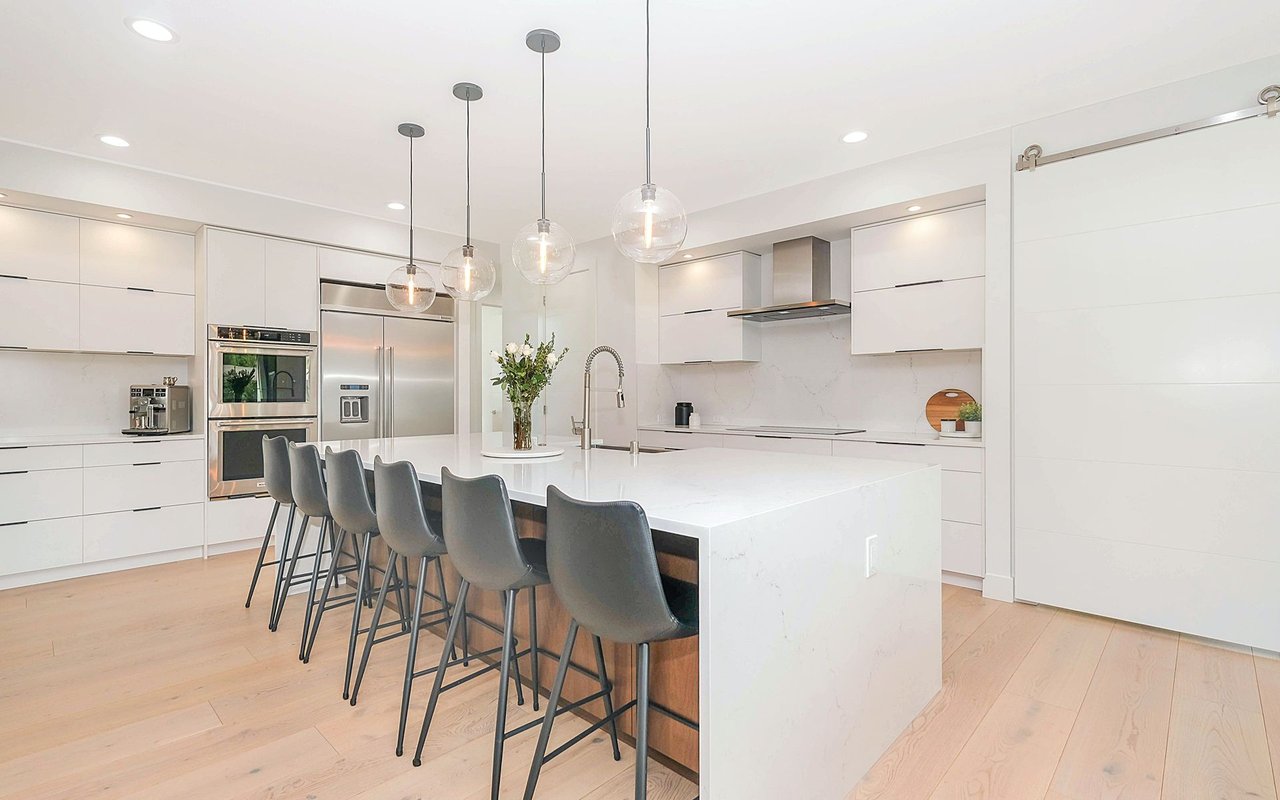 Modern white kitchen with a large island, gray bar stools, and stainless steel appliances.