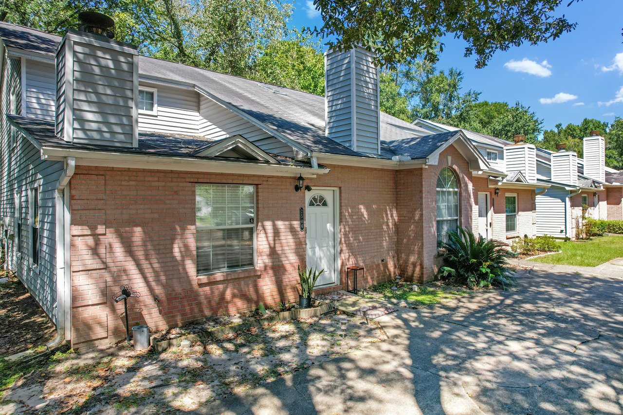 Angled view of the front exterior of a brick townhome at 2506 Nugget Lane in Tallahassee, Florida. The home features a white front door, large arched window, rectangular window, and two chimneys with gray siding. A concrete driveway and small landscaped area with plants and shrubs sit in front, with additional townhomes visible along the row.