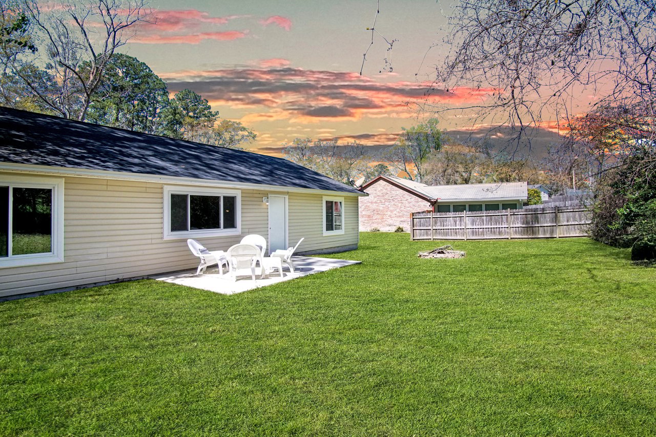 Backyard with green lawn, beige house, and white patio furniture under a vibrant sunset sky. Calm and inviting ambiance.