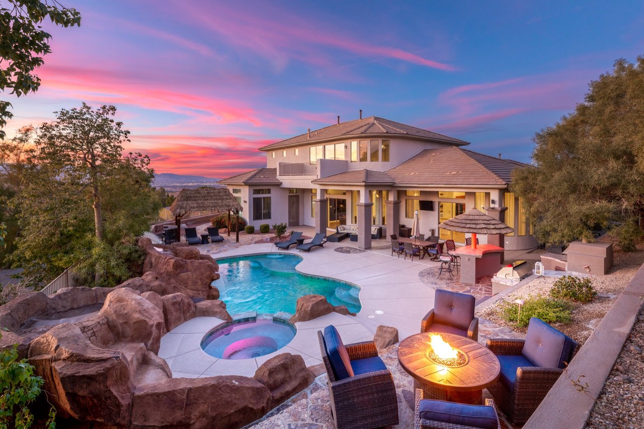 Luxury two-story home in Anthem Country Club, Henderson, Nevada at twilight, with a lagoon-style pool, spa, rock waterfall, and fire pit lounge under a pink sunset sky