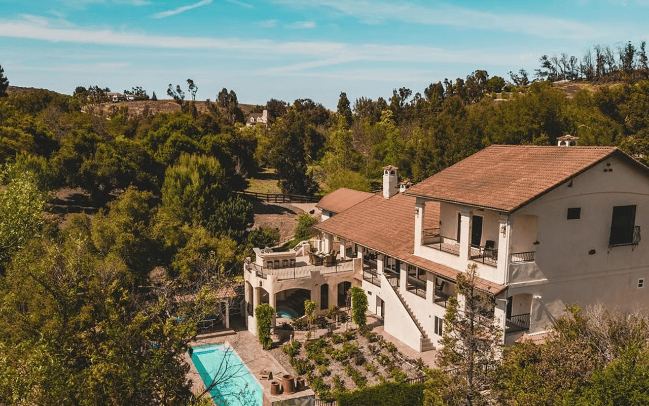 Hillside home with terracotta roofs, terraced balconies, and a turquoise pool amid wooded hills.