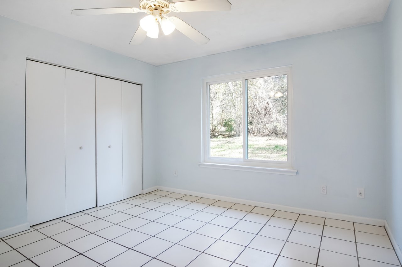 Empty, sunlit room with light blue walls and white tiled floor. A window on the right shows a garden. White folding closet doors and a ceiling fan.